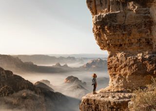 Person standing on a cliff edge above the clouds - Viking Travel