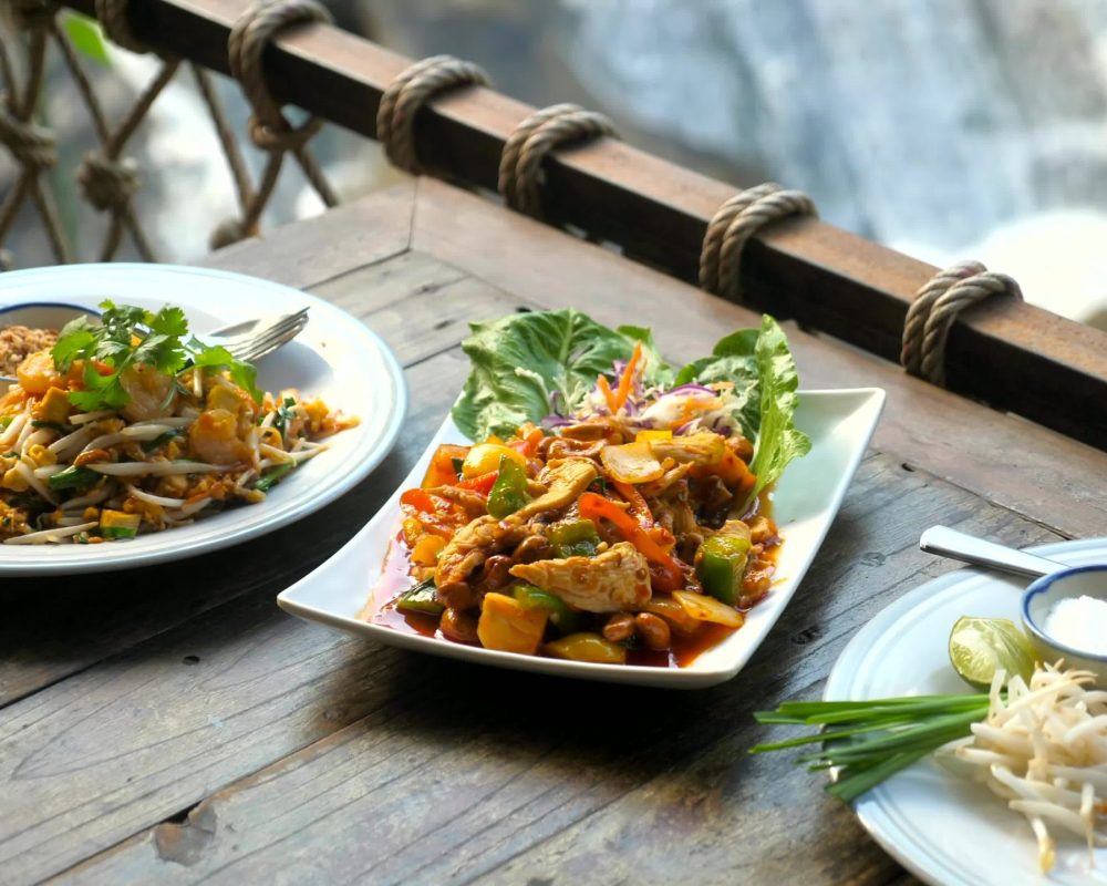 Traditional thai food in outdoor cafe with nature waterfall background. Fried noodle with prawn pad thai and chicken cashew nut on wooden table in restaurant. Asian style food lunch Traditional thai food in outdoor cafe with nature waterfall background. Fried noodle with prawn pad thai and chicken cashew nut on wooden table in restaurant. Asian style food lunch - Viking Travel