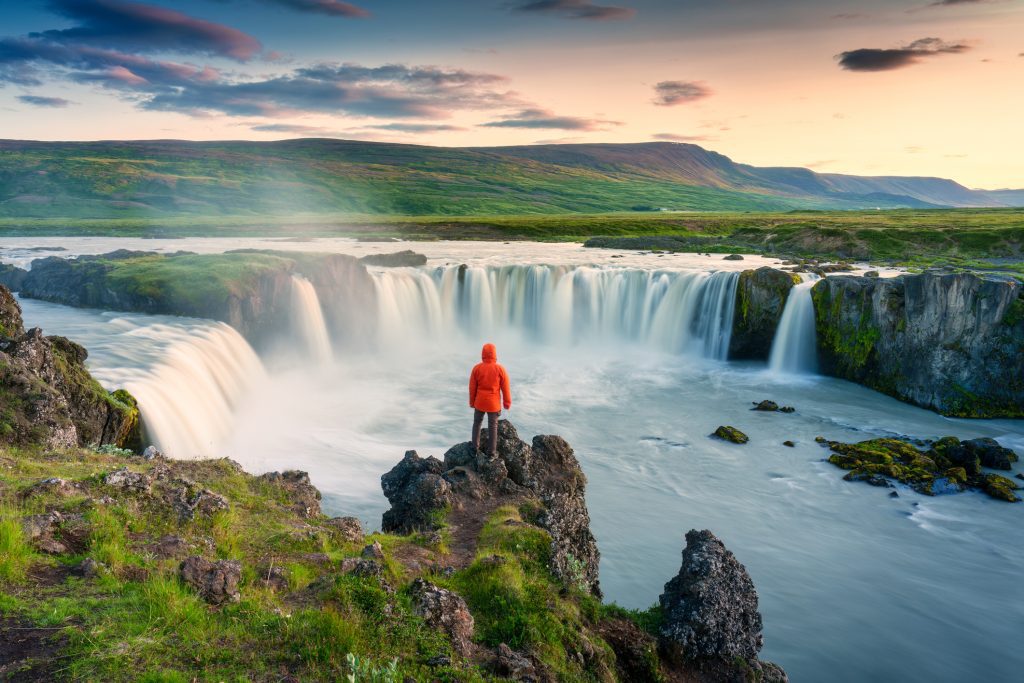 Godafoss waterfall flowing with colorful sunset sky and male tourist standing on cliff in summer at Iceland - Viking Travel